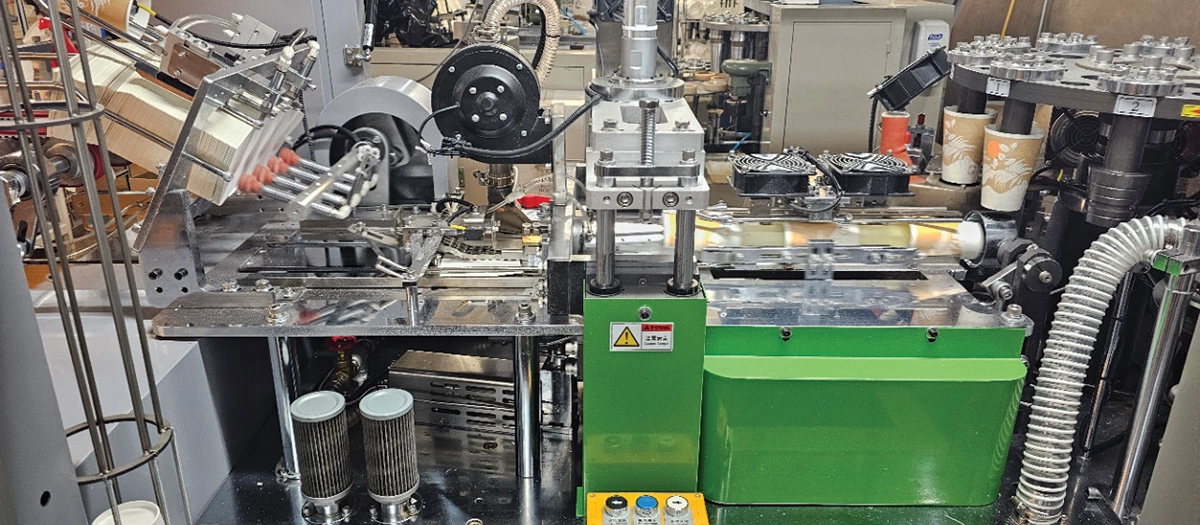 A wide, close-up shot of a complex industrial machine used for manufacturing paper coffee cups; The machine shows various mechanical stages, including a stack of flat paper blanks being fed on the left, a central green press unit with cooling fans, and a rotating carousel on the right where finished cups with printed designs are assembled