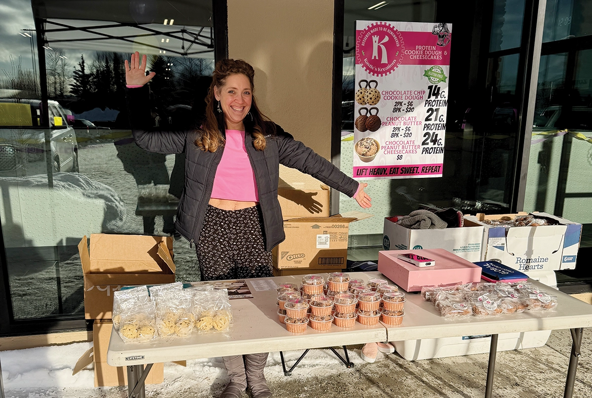A smiling woman in a black jacket and pink top stands behind a white folding table outdoors in a snowy setting; The table is filled with packaged baked goods, including cookie dough and cheesecakes; A pink sign titled Kastle's Kreations is posted on the window behind her, advertising protein-rich desserts