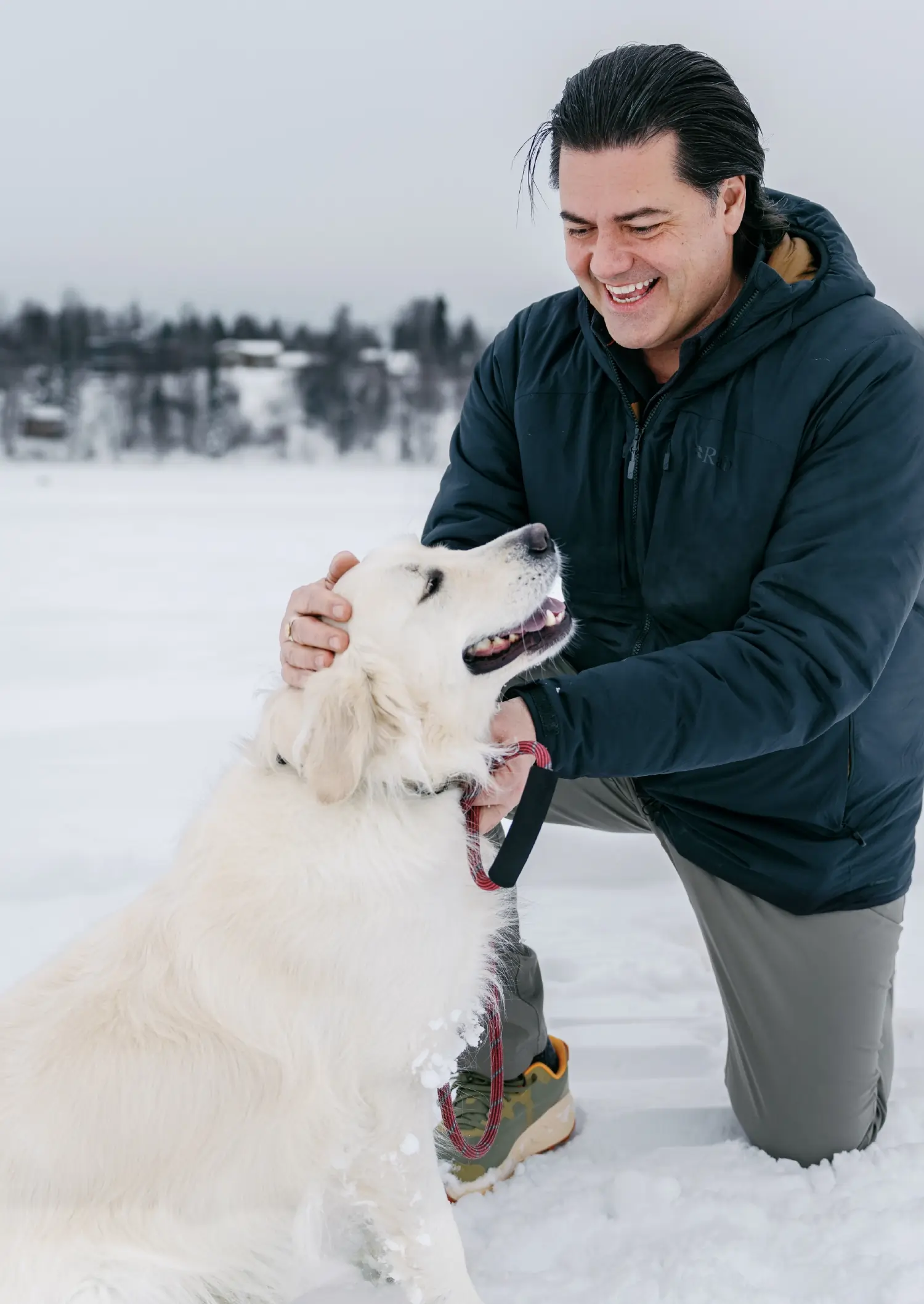 A candid outdoor shot of David McCarthy, a smiling man with dark hair, wearing a navy puffer jacket, grey trousers, and olive green fitness shoes kneeling in the snow to pet a large, white Golden Retriever; The background shows a snowy landscape with distant evergreen trees under a mostly overcast cloudy sky
