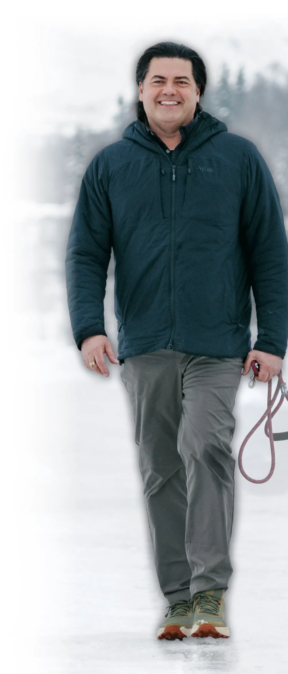 A full-length photograph of David McCarthy, a smiling man with dark hair, wearing a navy blue hooded puffer jacket, grey trousers, and olive green fitness shoes; He is walking through a snowy outdoor landscape and holding a red dog leash in his left hand