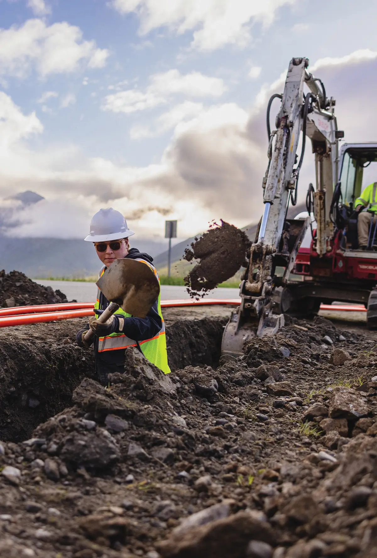 A worker in a hard hat and safety vest stands in a trench, using a shovel to move dirt. To the right, an excavator arm operates near orange telecommunications conduit. Mountains and a cloudy sky are visible in the background.
