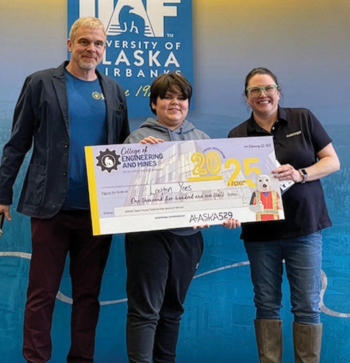 Two adults and a youth hold a large $1,500 ceremonial check in front of a University of Alaska Fairbanks backdrop. The check is awarded to Laughton Yoes for the Engineering Open House Scholarship and is sponsored by Alaska 529.