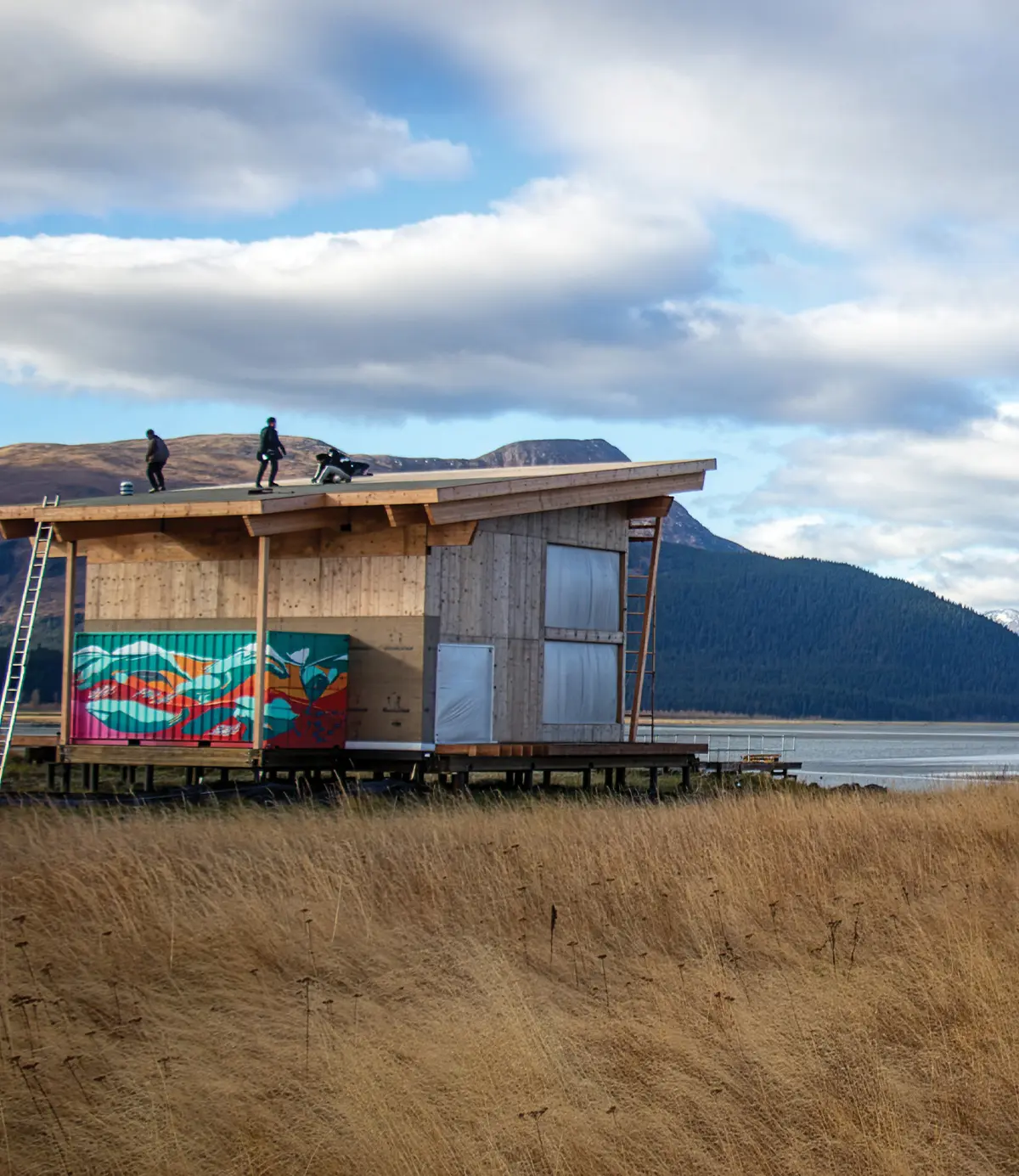 A wooden building under construction on stilts overlooks the water, with a mural-painted shipping container integrated into the base. Three people stand on the roof. Dry grass and mountains are in the background.