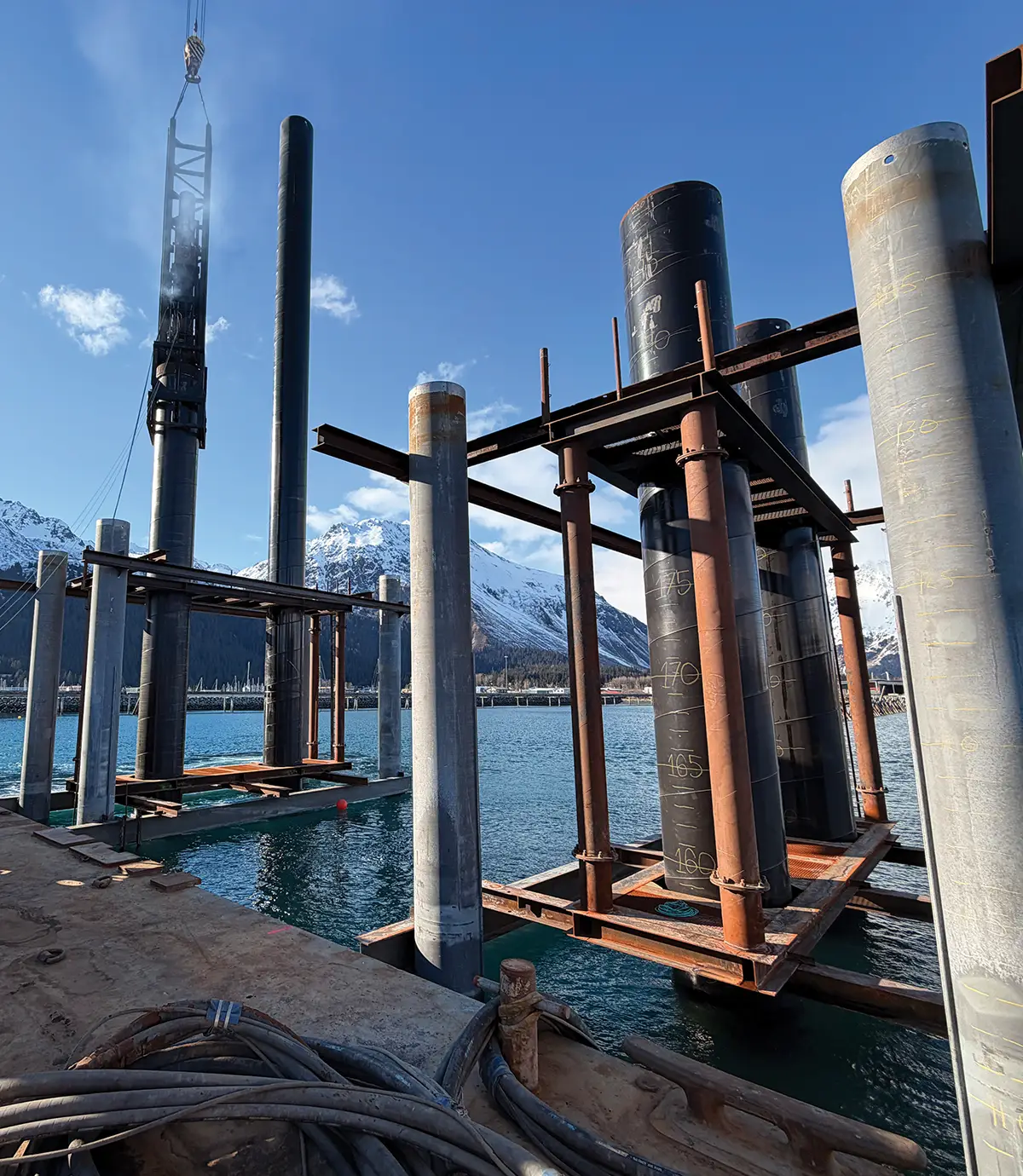 Large metal pilings and steel support frames stand in the water at the Port of Seward during construction. A crane is visible to the left, and snow-capped mountains rise in the background under a clear sky.