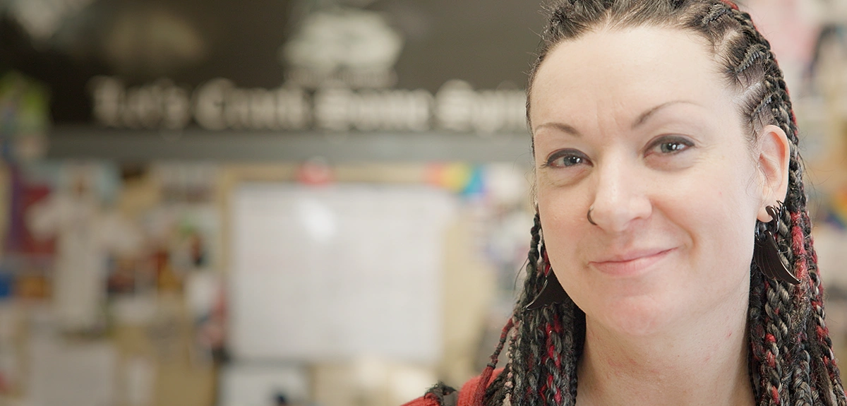 Taylor Jordan, owner of Black Birch Books, smiles in a close-up portrait. She has dark hair in braids with red highlights and wears wing-shaped earrings. Behind her is the blurred interior of a bookstore and workshop space.