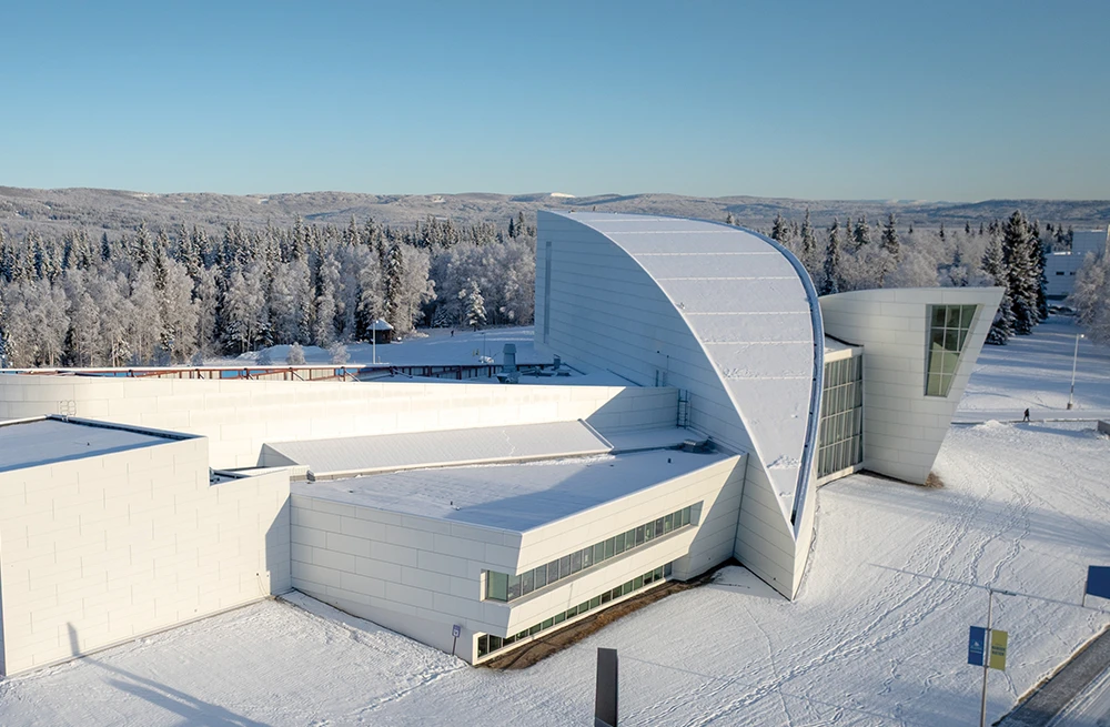 An aerial view of the snow-covered University of Alaska Museum of the North and its new planetarium addition. The modern white building sits among snowy fields and a dense forest of frosted trees.