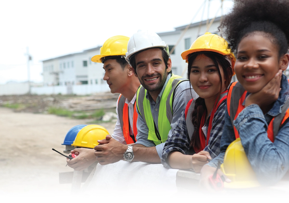 Four diverse workers in hard hats and safety vests lean over building plans at an outdoor construction site. Two look toward the camera while others look ahead. Buildings under construction are visible in the background.