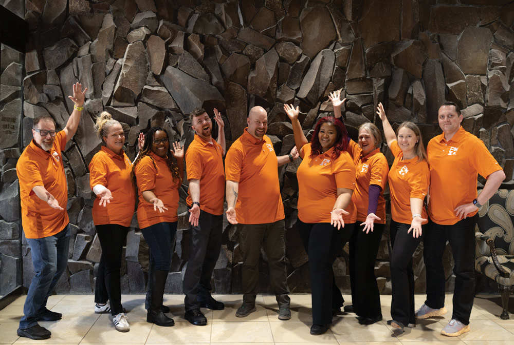 A diverse group of nine people wearing matching bright orange polo shirts pose playfully in front of a dark, textured stone wall. They are smiling with their arms raised or gesturing forward.