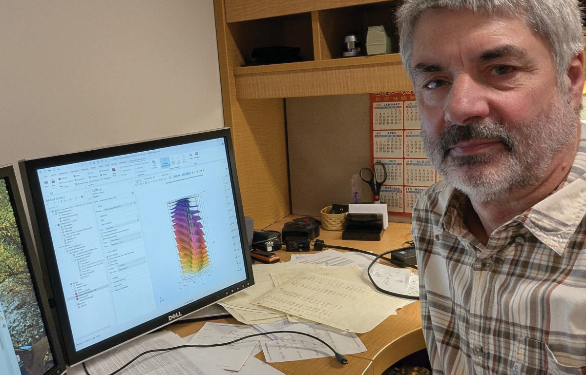 A man with a grey beard sits at a desk with a computer monitor displaying complex 3D data models and scattered research papers.