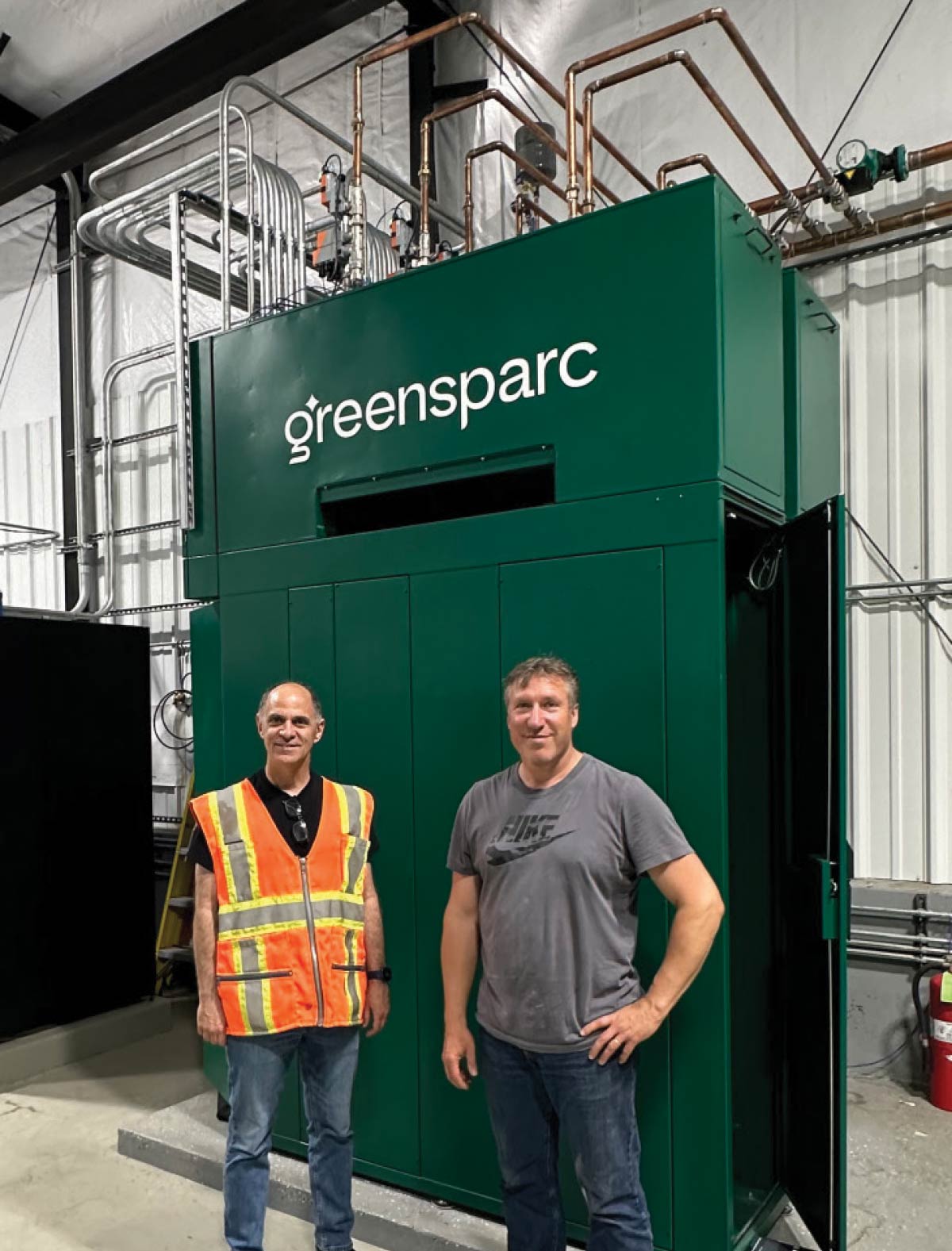 Two men stand in front of a large, green industrial unit labeled "greensparc" inside a facility with exposed pipes and conduits.