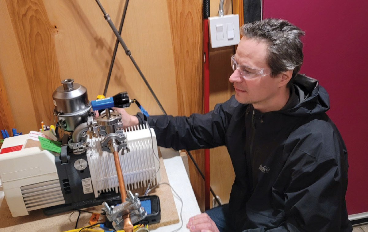 A man wearing safety glasses carefully inspects and adjusts a piece of scientific laboratory equipment with various tubes and wires.