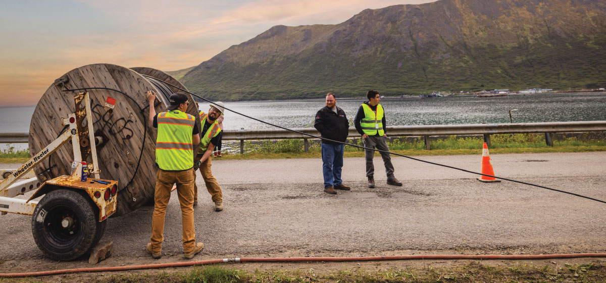 Workers in safety vests manage a large wooden cable reel on a roadside, with a body of water and rolling hills in the scenic background.
