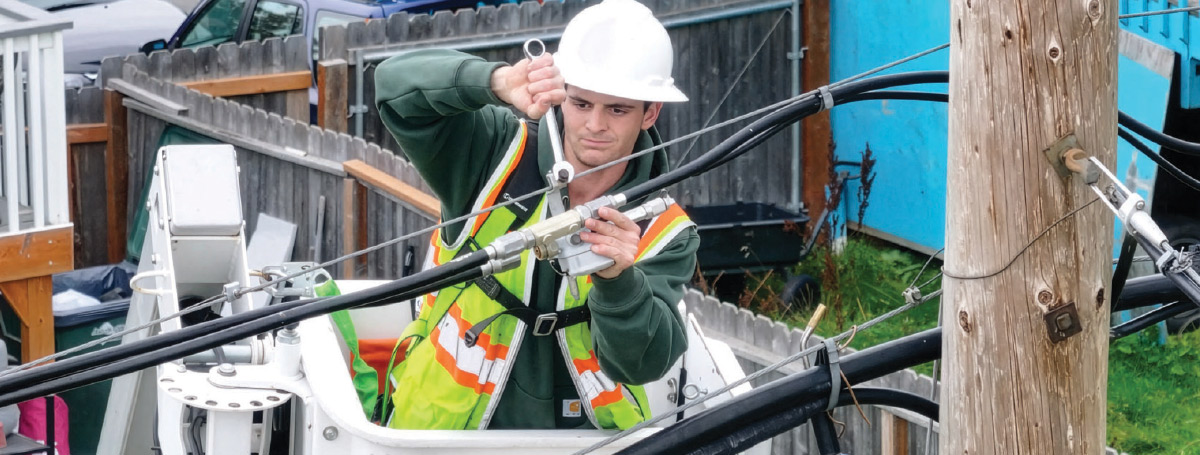 A utility worker in a hard hat and high-visibility vest uses a wrench to work on overhead cables from a white aerial lift bucket.
