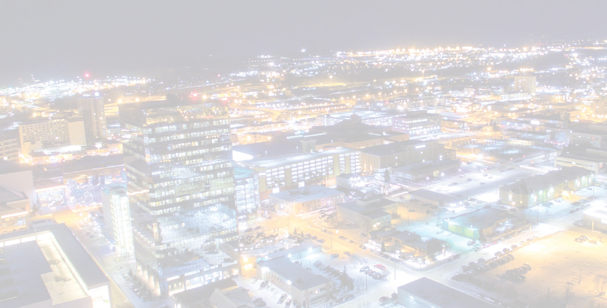 An aerial, high-key nighttime view of a brightly lit city skyline with glowing office buildings and streetlights under a hazy sky.