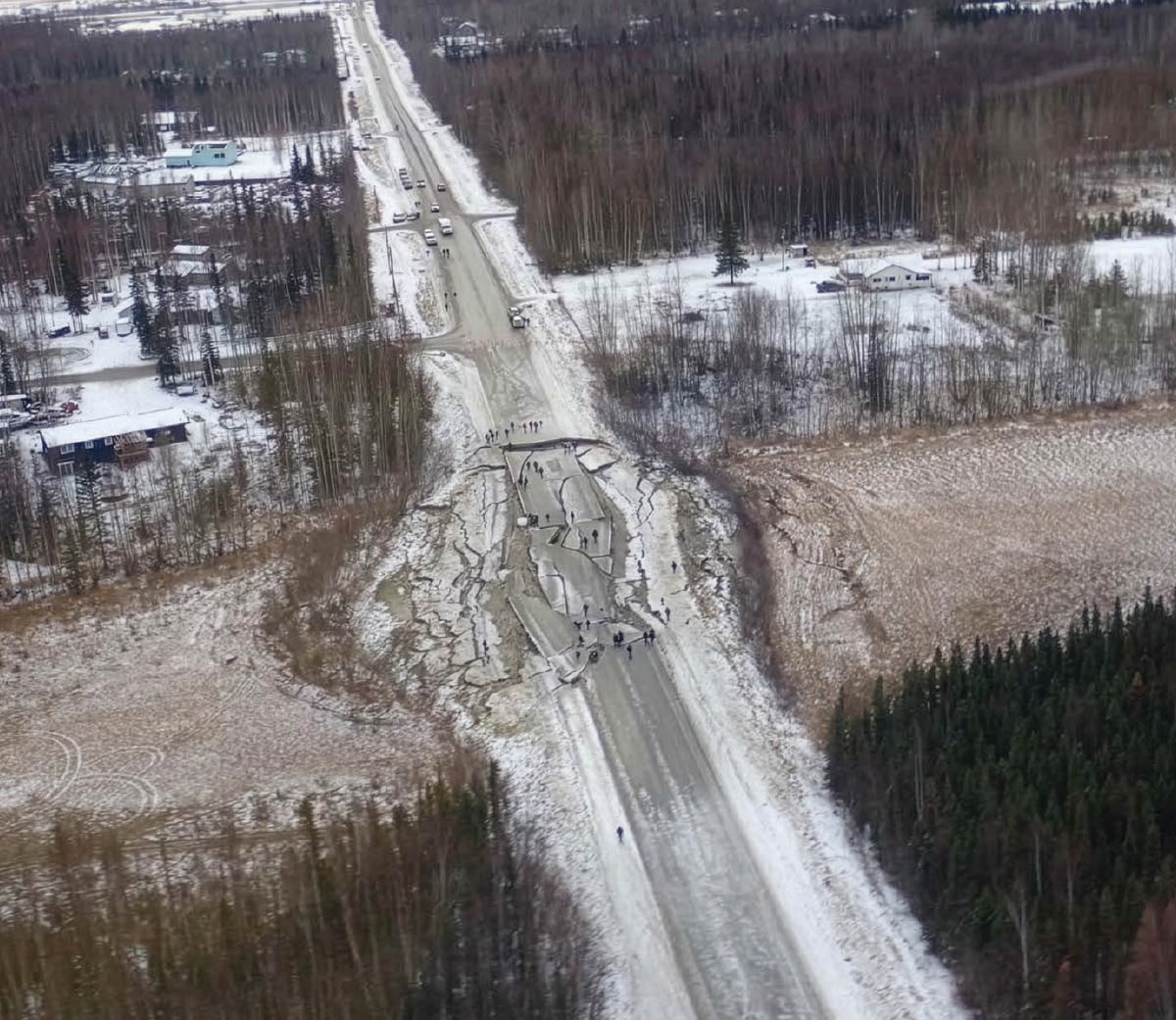 An aerial view of a long, snowy road showing severe earthquake damage, with large cracks and collapsed sections of asphalt blocking traffic.