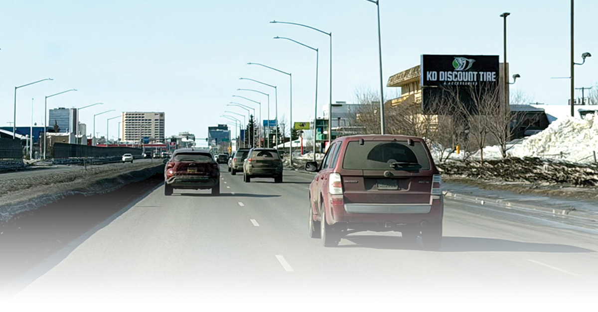 Vehicles traveling on a multi-lane urban highway toward a city skyline. A "KD Discount Tire" sign stands on the right, and snow banks line the road under a clear sky.