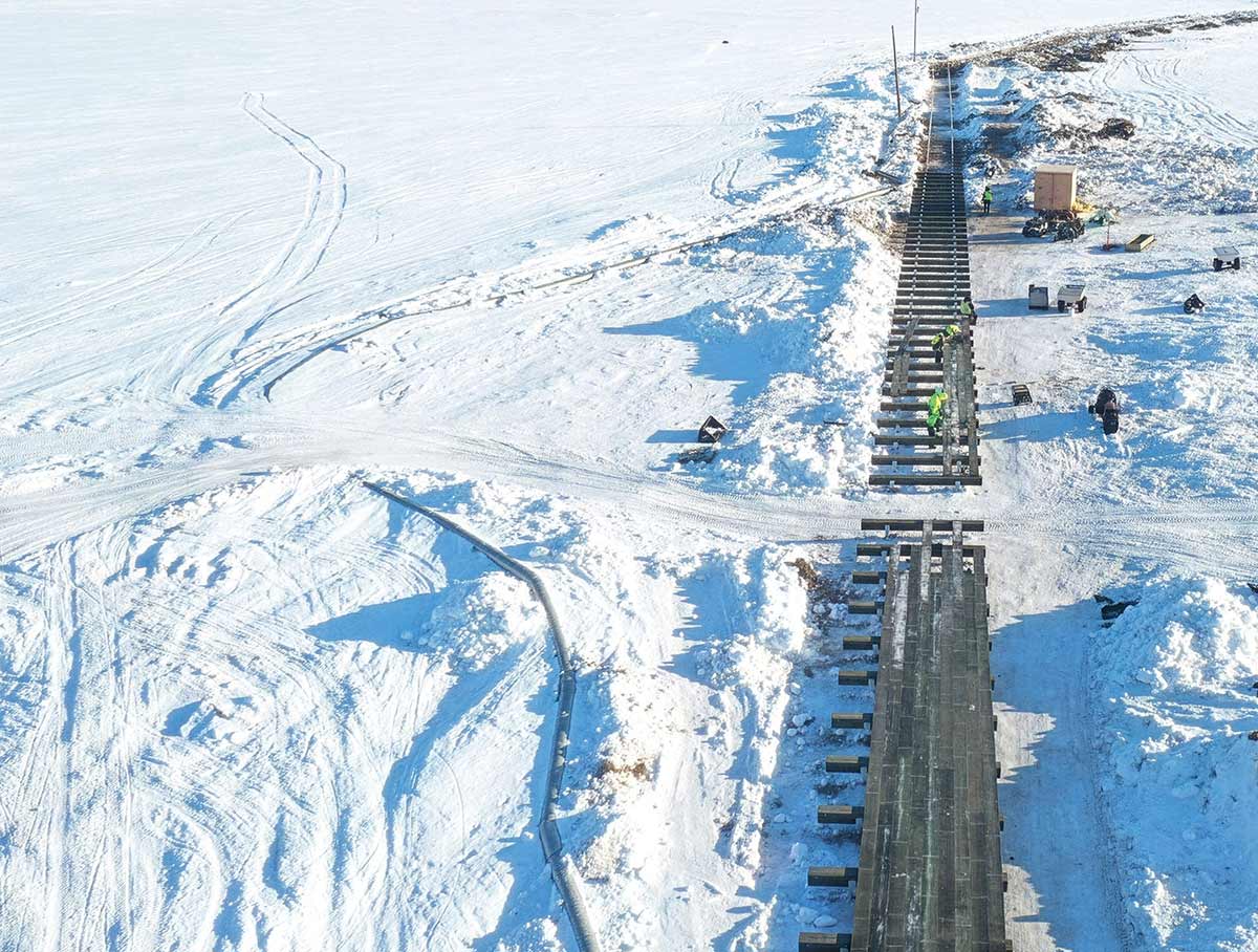An aerial view of a construction crew working on a long wooden pier or boardwalk in a vast, bright, snow-covered landscape
