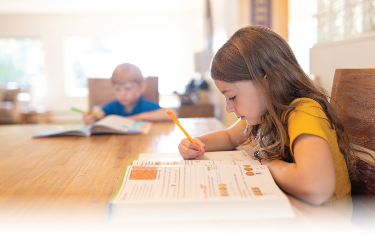 A young girl focuses on writing in a workbook at a wooden table, while a young boy studies in the soft-focus background.