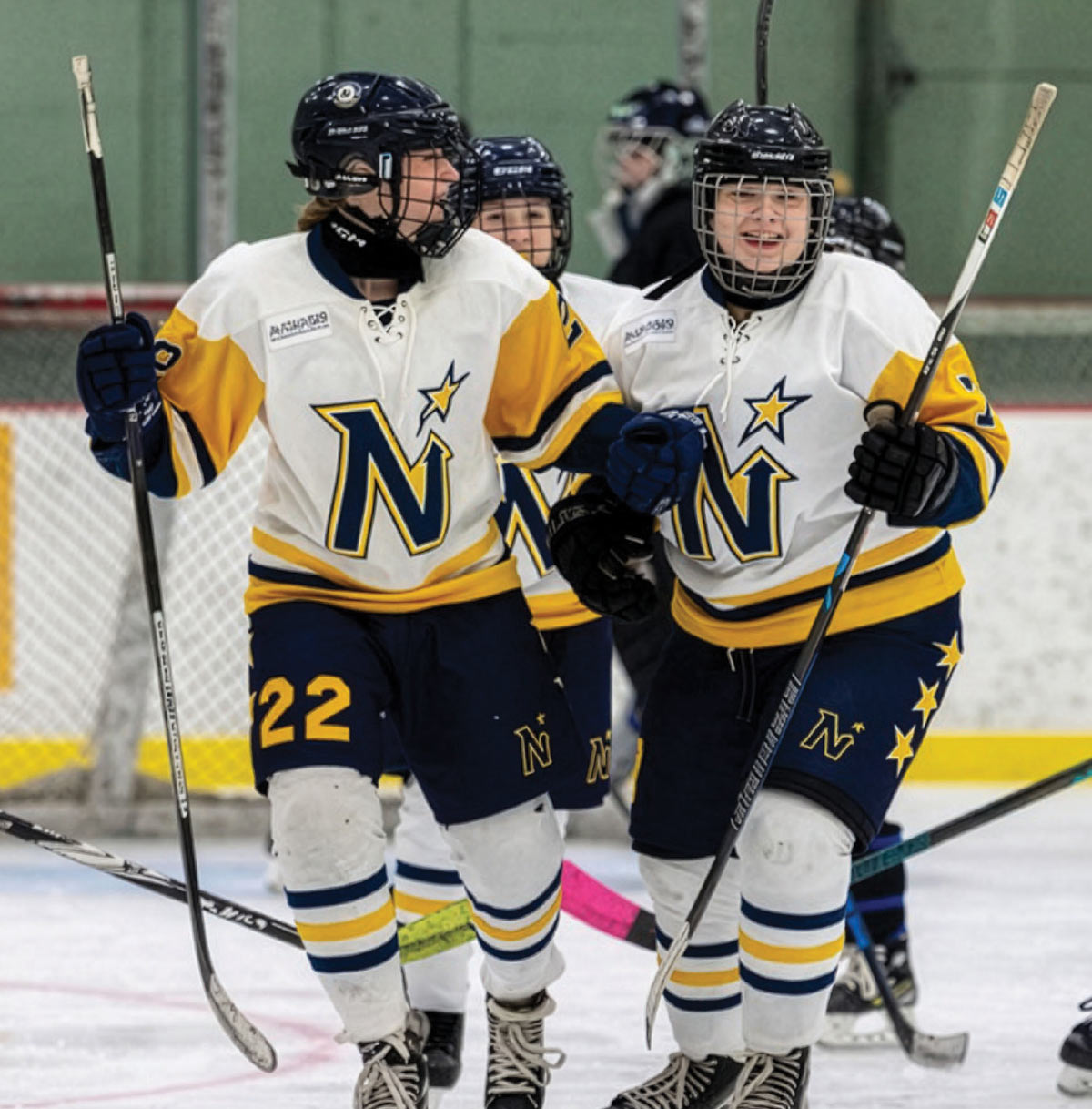 Two youth ice hockey players in white, blue, and gold uniforms celebrate on the ice, smiling and holding their hockey sticks.