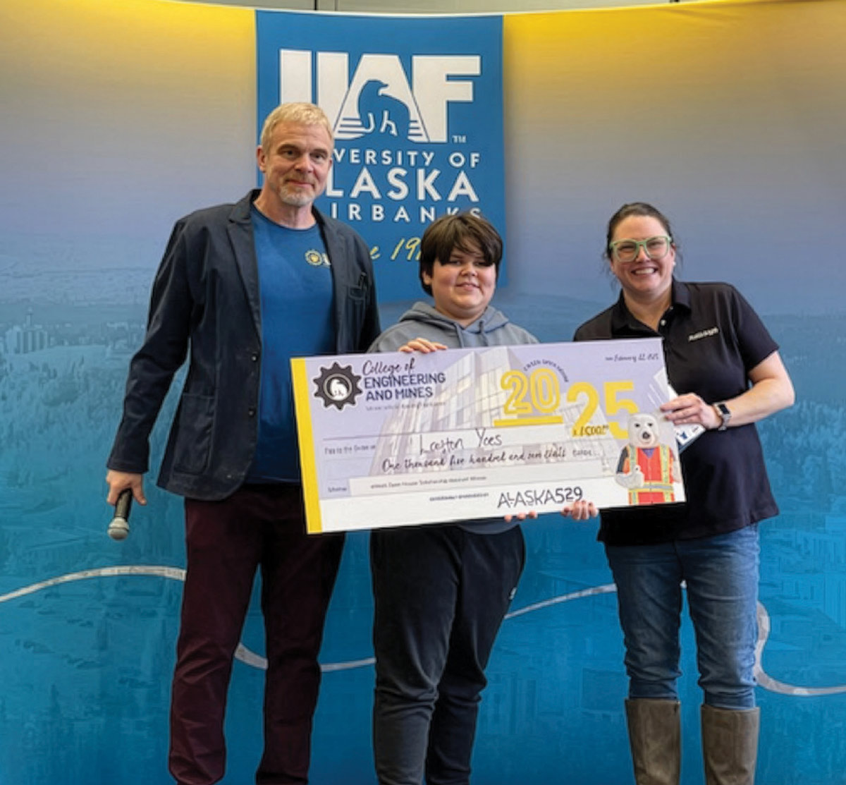 A student holds a large scholarship check alongside two adults in front of a University of Alaska Fairbanks backdrop.