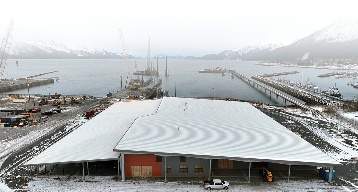 Aerial view of a modern industrial building at a snowy port. Marine construction cranes and docks are visible in the water, with snow-covered mountains in the distance under a grey sky.