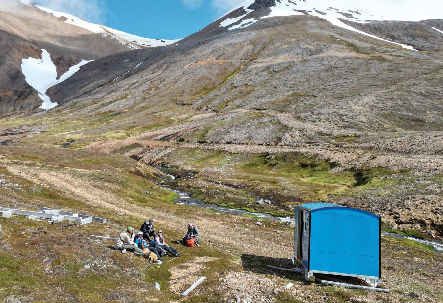 A geological research site in a mountainous tundra region with five researchers sitting on the grassy ground nearby a bright blue portable shelter