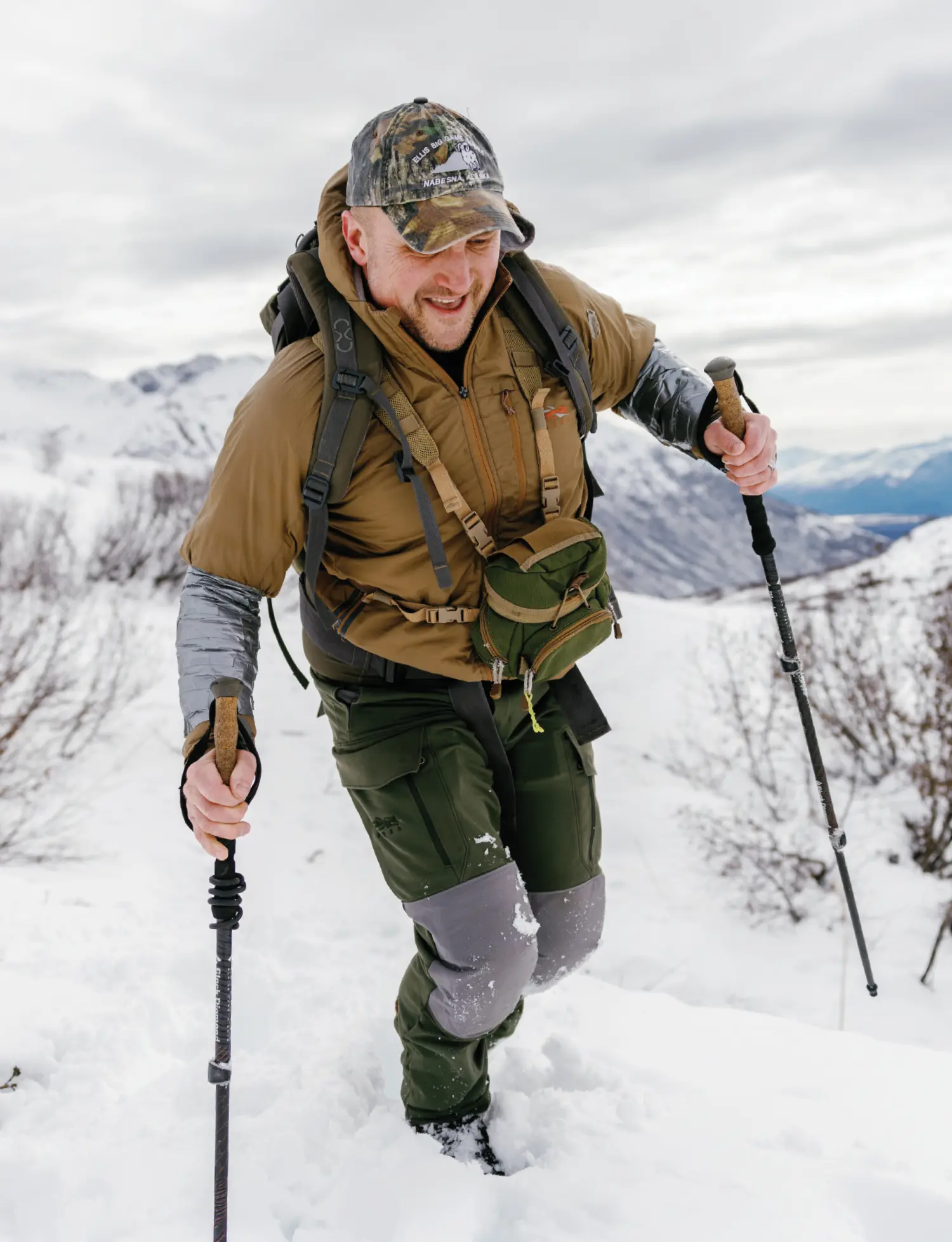 Jeff Miller, a man hiking uphill with trekking poles through deep snow on a cloudy day; He is outfitted in professional hiking outdoor gear attire
