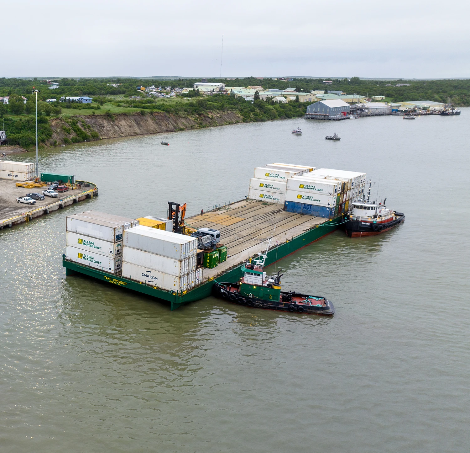 Aerial drone shot of a green and tan flat-top cargo barge, the Taku Provider, being maneuvered by two tugboats on a river
