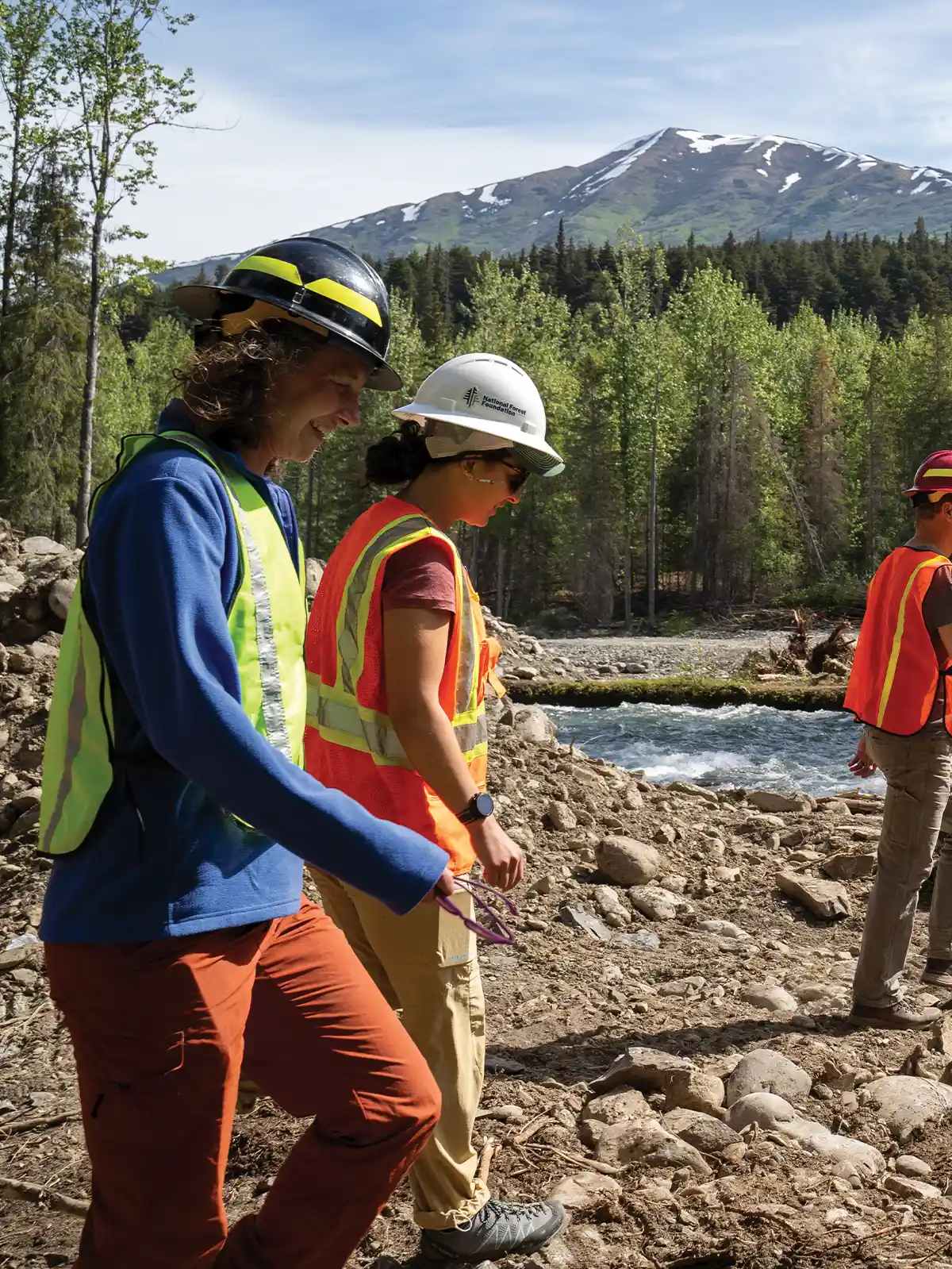 Two workers in hard hats and safety vests walk along a rocky riverbank. A fast-flowing river, dense forest, and snow-capped mountain are in the background.