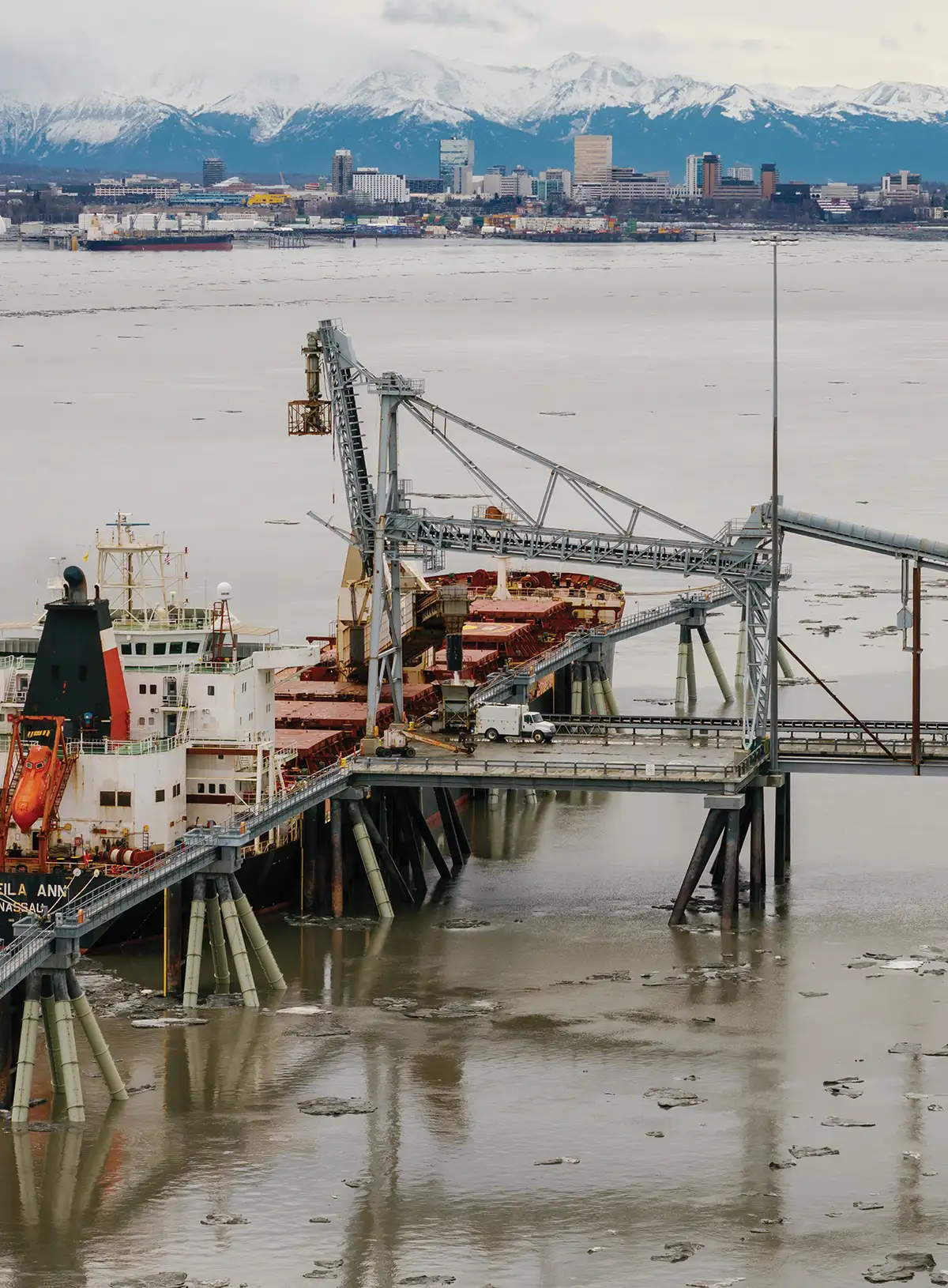 A large cargo ship docked at an industrial port with loading cranes. In the background, a city skyline sits before a range of snow-covered mountains. Ice floes dot the water.