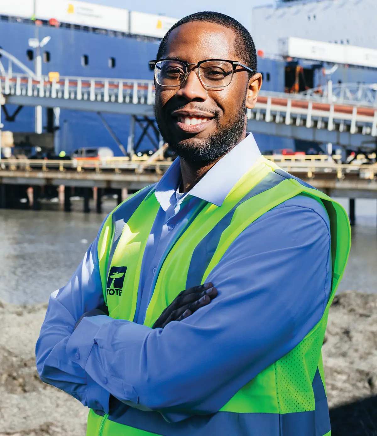 Portrait of a smiling man with glasses wearing a neon safety vest with the TOTE logo. He stands outdoors in front of a large blue cargo ship at a port.