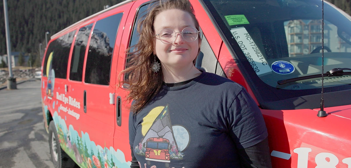 A woman in glasses stands in front of a red "Red Eye Rides" transport van. The van features a landscape mural and text indicating service between Seward and Anchorage.