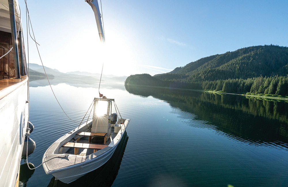 A metal skiff is lowered from a larger vessel into calm, reflective water. The sun shines brightly over a lush, forested shoreline in the background.