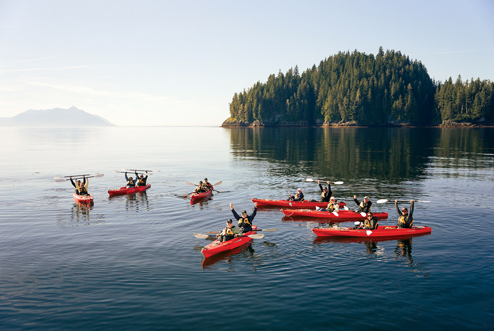 A group of people in red double kayaks on calm water raise their paddles and hands in celebration. A densely forested island and distant mountains are in the background.