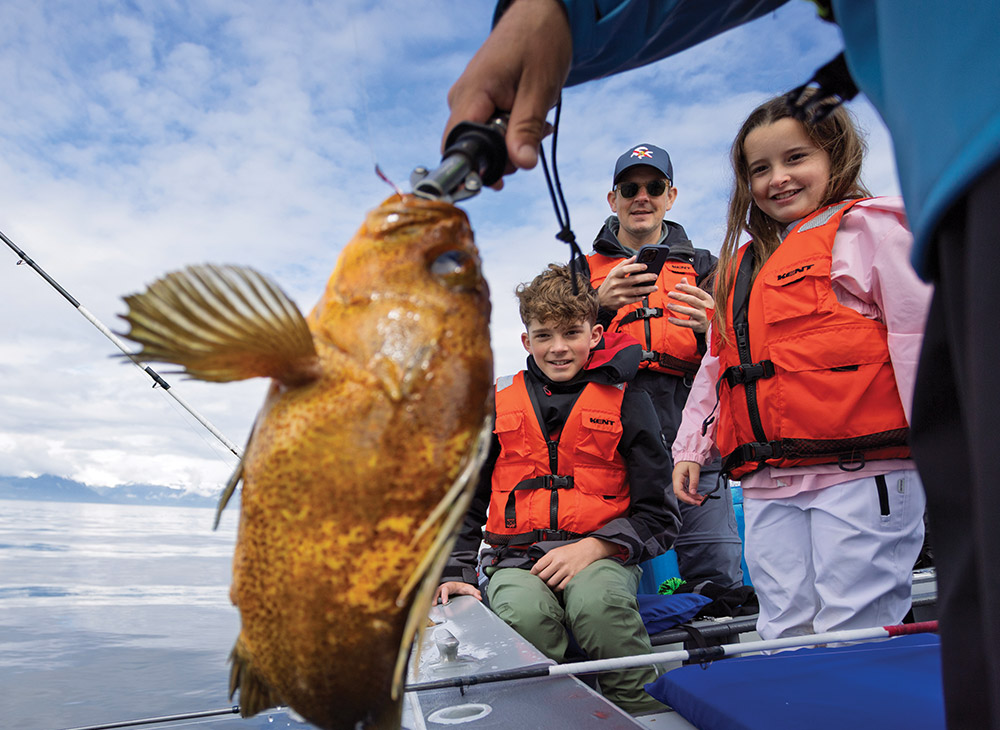 A close-up of a large, mottled orange-brown rockfish held up on a fishing line. In the background, a man and two children wearing orange life vests watch from a boat.