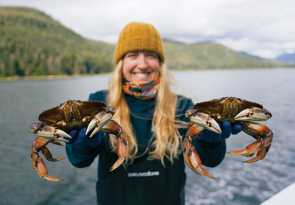 A smiling woman in a yellow beanie and waterproof bibs holds up two large crabs. A body of water and forested mountains are in the background.