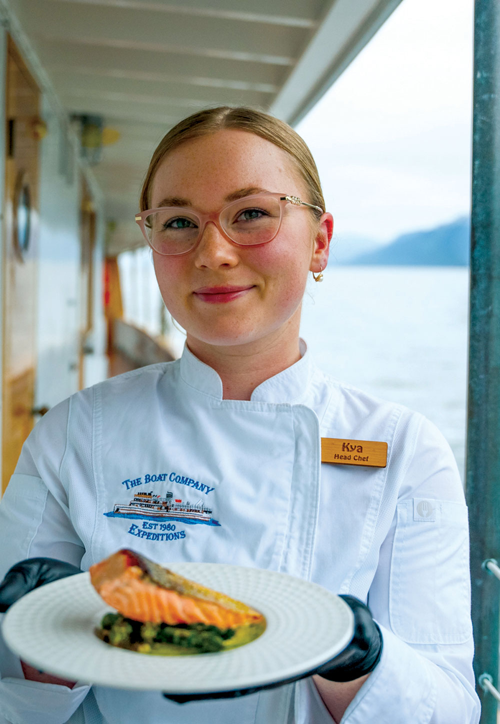 A head chef in a white uniform holding a plate with a grilled salmon fillet served over green vegetables. The background shows the deck of a boat and misty mountains.