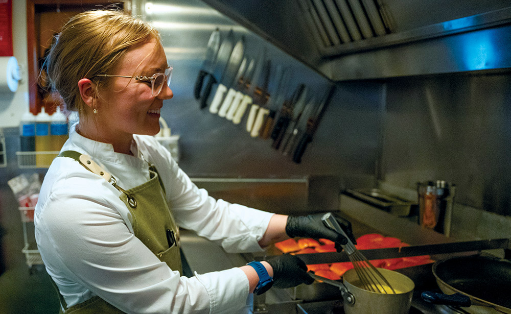 A chef in a kitchen wearing an apron and glasses whisks a sauce in a small pot on a stove. Professional knives are mounted on the wall behind her.