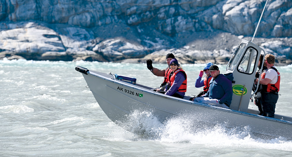 A small motorboat carrying five people moves through choppy water near a rocky cliff. One person waves toward the camera.