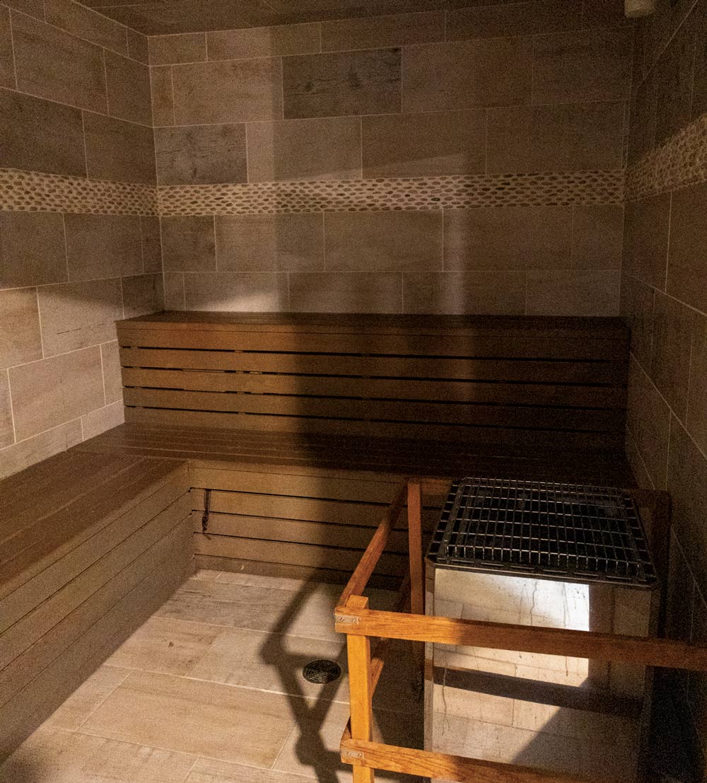 Interior of a wooden sauna with tiered bench seating, stone tile walls, and a metal heating unit.