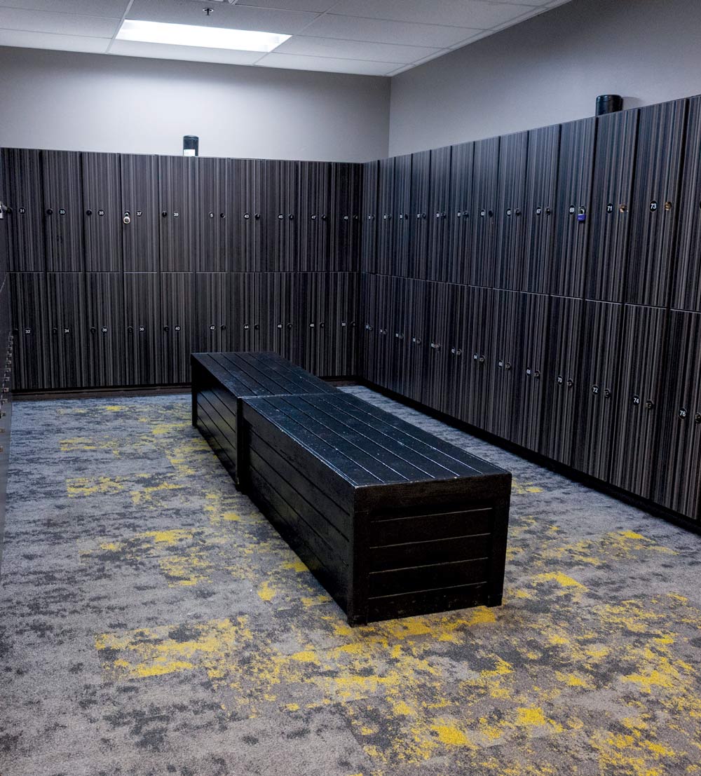Gym locker room with dark wood-grain lockers, a central black bench, and grey and yellow patterned carpet.