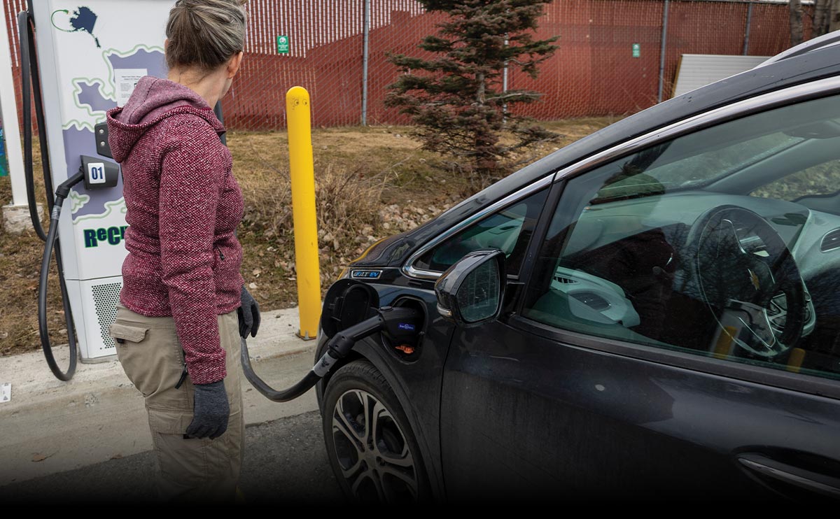 A woman in a maroon sweater charges a dark grey Chevrolet Bolt EV at a white "Recharge" station featuring a map of Alaska. The charging cable is connected to the car's front port in an outdoor parking area.