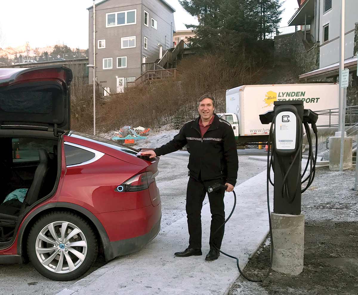 A smiling man holds a charging cable next to a red Tesla Model X with its hatch and wing door open. The snowy outdoor scene includes an EV charging station, a Lynden Transport truck, and a grey building on a hill.
