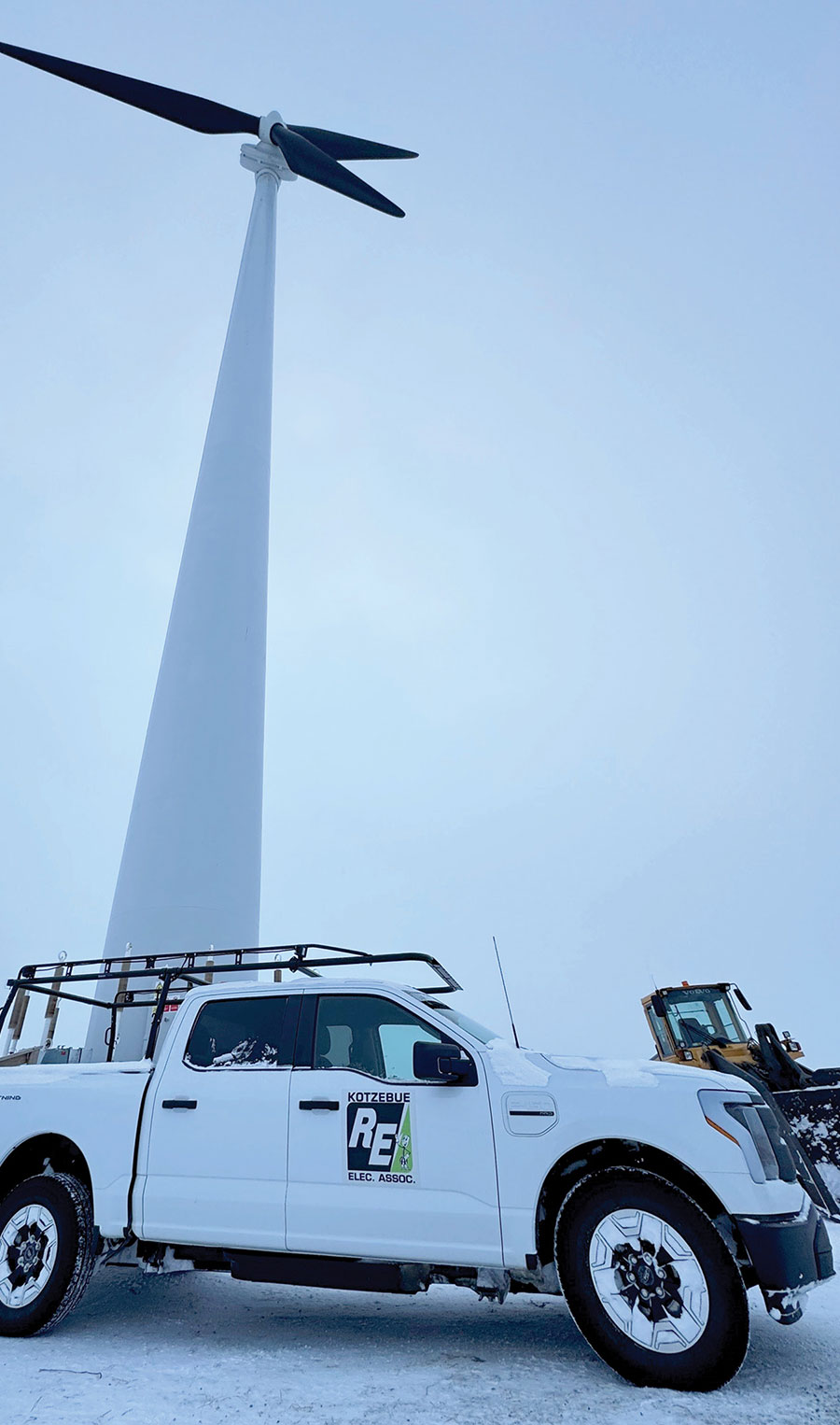 A white Ford F-150 Lightning truck with the Kotzebue Electric Association logo parked on snow beneath a towering wind turbine. A yellow loader is visible in the background against an overcast sky.