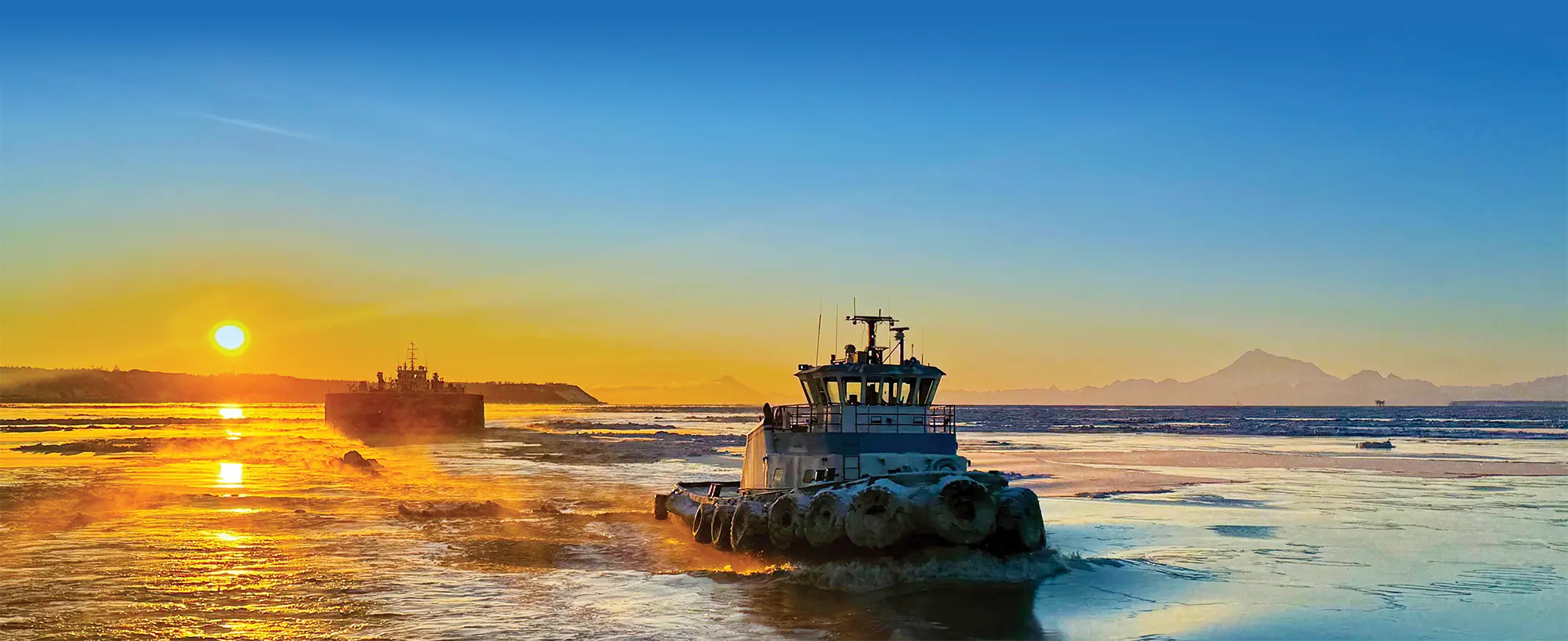 A tugboat and industrial ship navigate icy waters during a vibrant golden sunset. Snow-capped mountains are visible in the distance under a blue and orange sky.