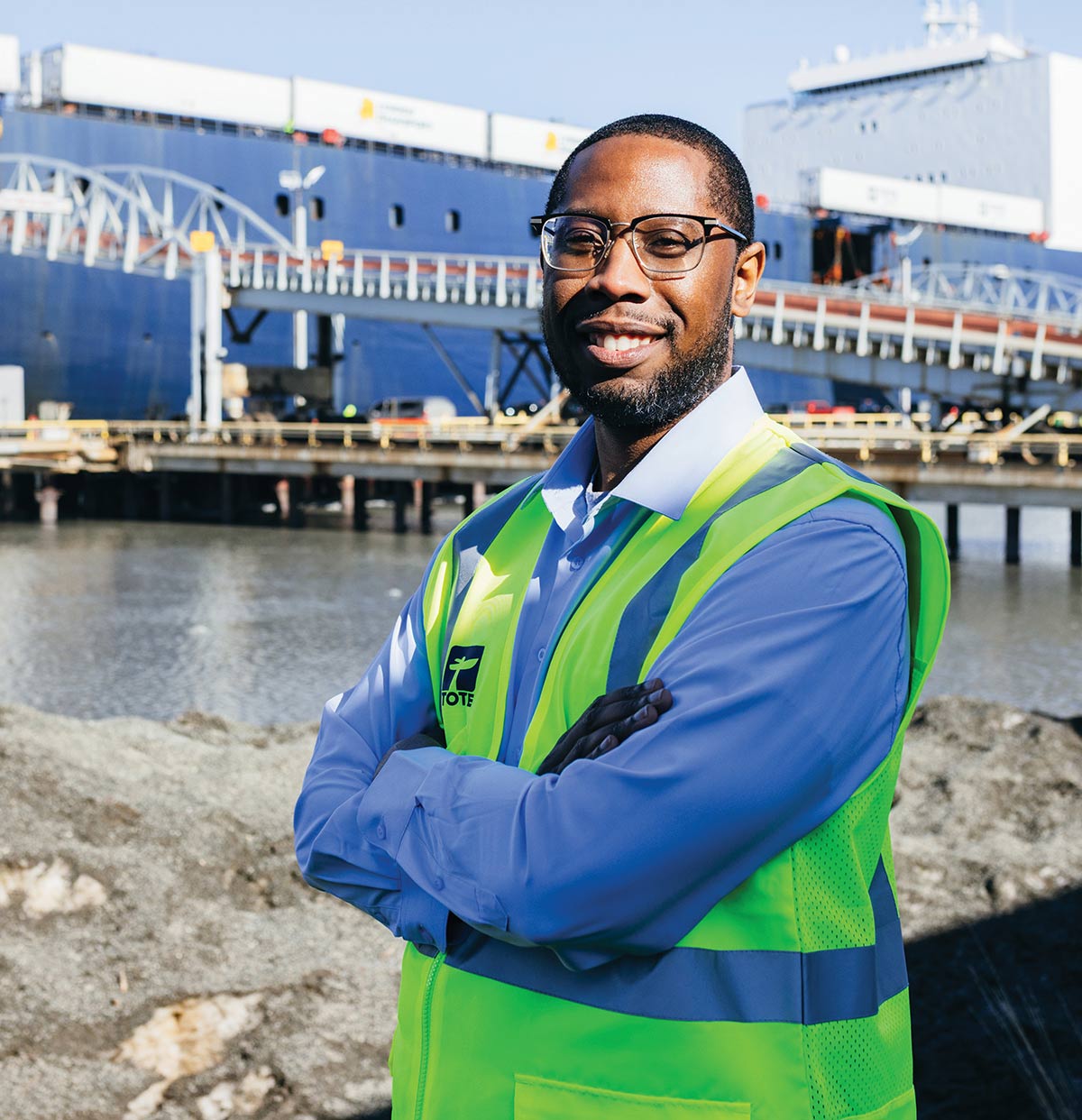 A smiling man in a neon green TOTE safety vest stands with arms crossed at a shipping port. A large blue cargo ship and a loading bridge are visible in the background under a clear sky.