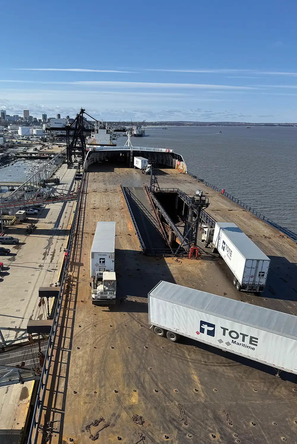High-angle view of a cargo ship deck at a port being loaded with TOTE Maritime semi-trailers. A city skyline and calm water are visible in the background under a clear blue sky.