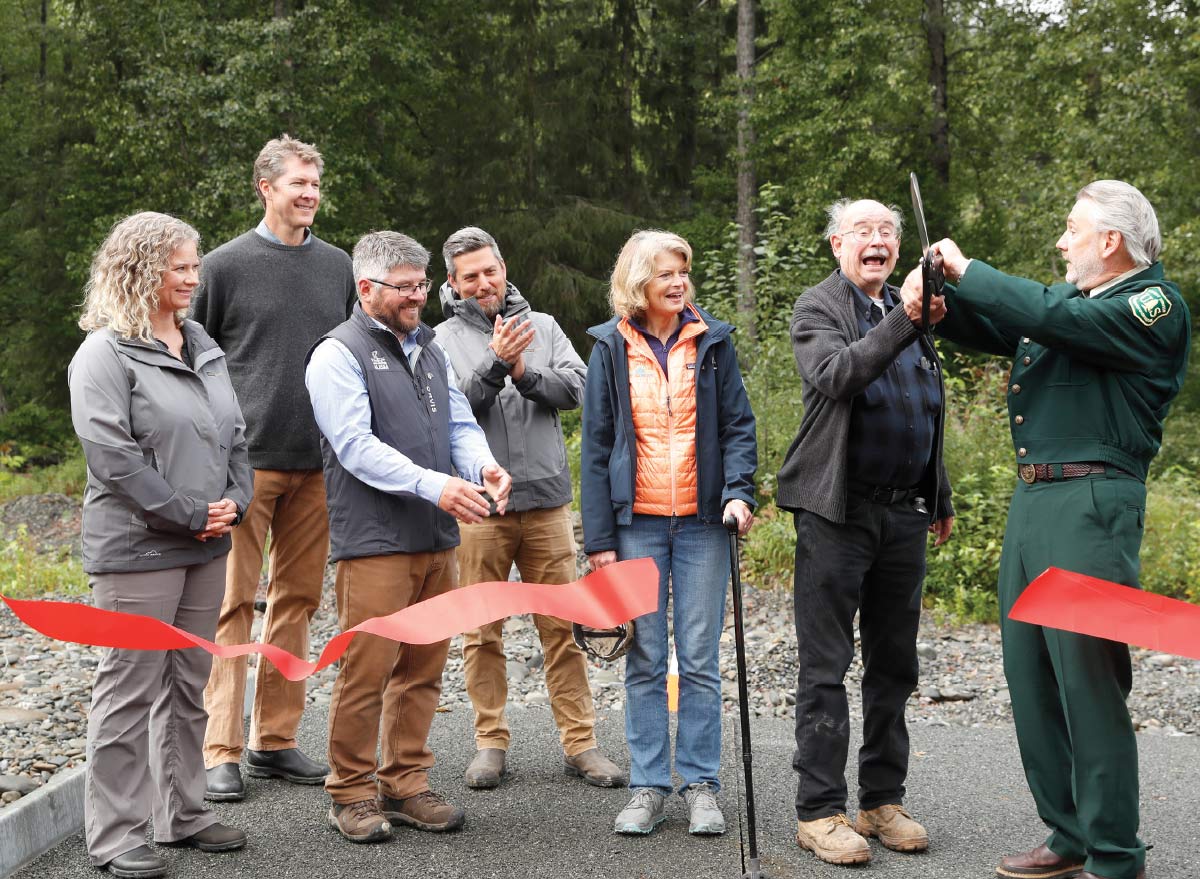Officials and community members celebrate with a ribbon-cutting ceremony outdoors, led by a man in a Forest Service uniform.