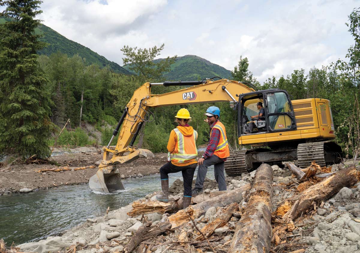 Two workers in safety vests and hard hats oversee a yellow excavator moving logs and earth near a stream in a forested mountain area.