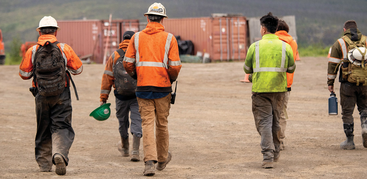 A group of workers in high-visibility gear and hard hats walk across a dirt site.